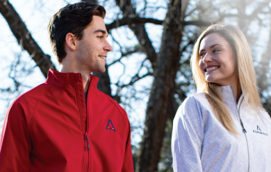 A woman in a white FEATHERLIGHT SOFT SHELL JACKET and a man in a red jacket stand outdoors, smiling at each other, with bare trees behind them.