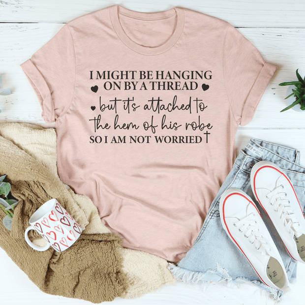 A pink Hanging On By A Thread T-Shirt made from 100% ring-spun cotton with "I might be hanging on by a thread…" text is displayed beside denim shorts, white sneakers, a heart mug, and a green blanket.